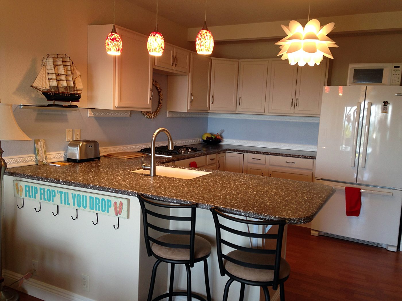 A kitchen with white cabinets, three glass pendant lights and a flower-shaped light over a speckled counter with two bar stools.