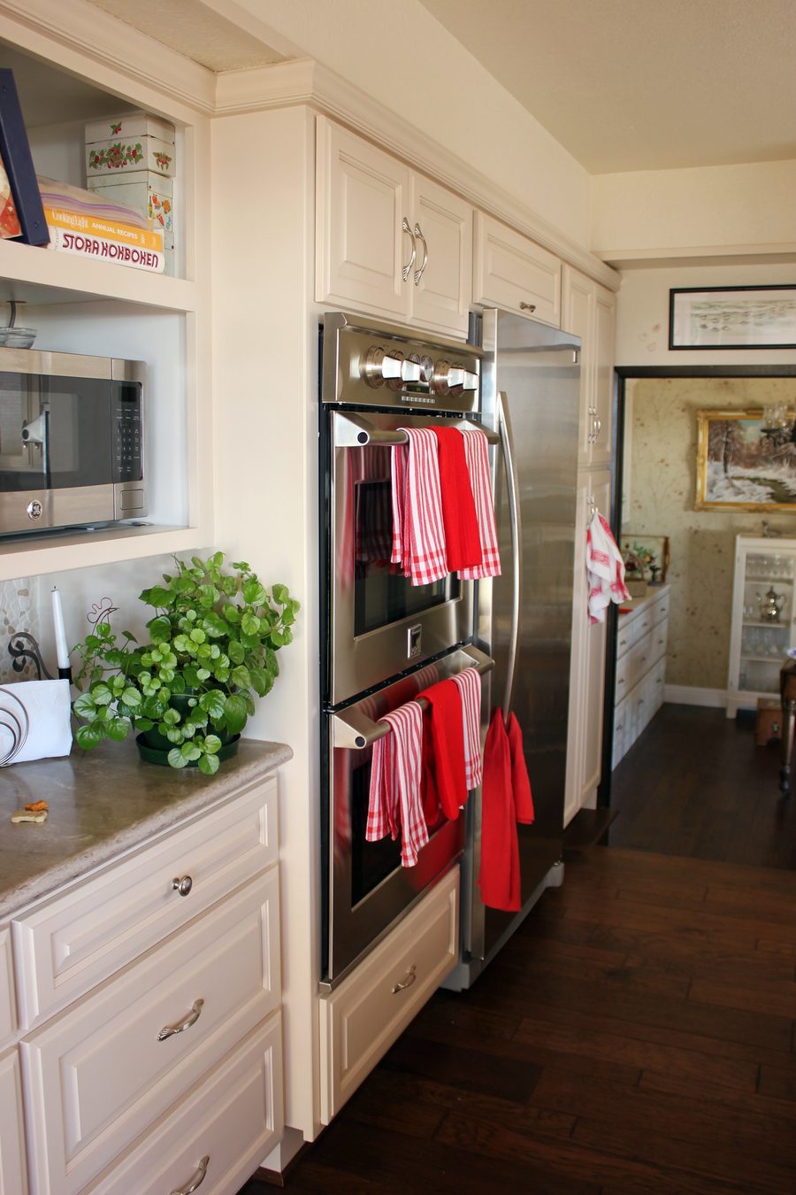 A kitchen wall of cream cabinets around stainless double wall ovens and a refrigerator, with red towels on the oven handles.