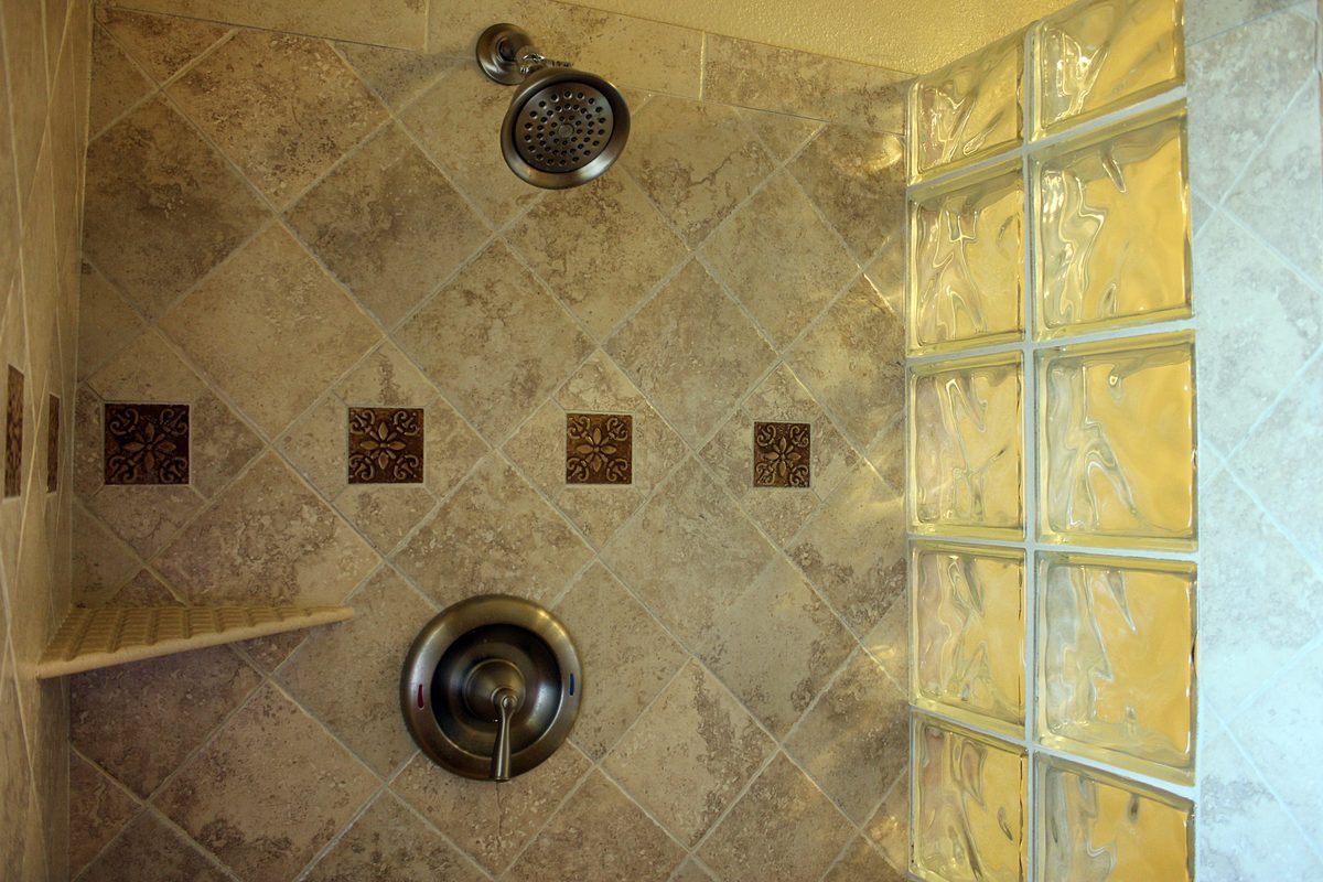 A tiled shower with diagonal stone-look tile, a row of small bronze accent tiles and a glass-block wall.